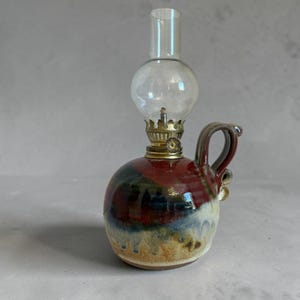 May include: A small, round oil lamp with a clear glass globe and chimney. The ceramic base features a handle and is painted with a blend of red, blue, and yellow hues. The lamp has a brass fitting.