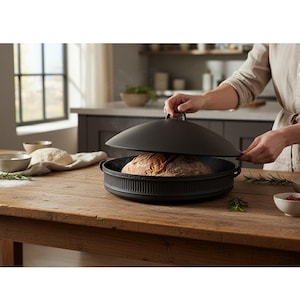 May include: A black cast iron bread baker with a domed lid, revealing a freshly baked loaf of bread. The baker sits on a wooden table, with steam rising from the bread. Rosemary and small bowls are also on the table.