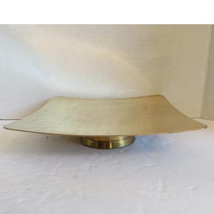 May include: A square brass tray with a slightly curved surface and a textured finish, supported by a circular pedestal base. The metal has a polished gold-toned appearance with subtle indentations across the flat top surface area.