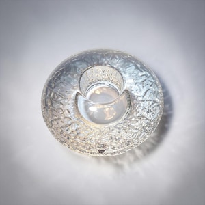 May include: A clear glass candle holder with a textured, leaf-like pattern. The candle holder has a circular base with a central well for the candle and a curved, crescent-shaped opening. The Orrefors Sweden logo is visible.