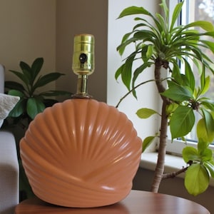May include: A peach-colored ceramic table lamp with a shell-like design. The lamp has a brass-colored socket and a round base. The lamp is sitting on a wooden table. A green plant is in the background.