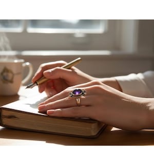 May include: A gold-toned ring with a large oval purple gemstone is worn on a hand writing in a notebook with a gold pen. The ring has a decorative setting. A cup of coffee sits nearby.