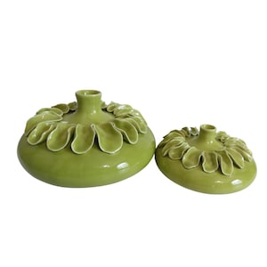 May include: Two green ceramic vases with a glossy finish. The vases have a unique design, with a ring of leaf-shaped embellishments around the top. The larger vase is round, while the smaller one is flatter. Both have a small opening at the top.