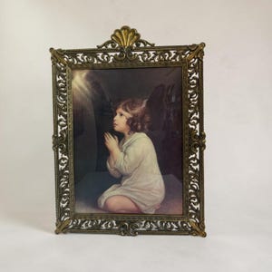 May include: A rectangular brass picture frame with an ornate, filigree scrollwork border and a shell-shaped crest at the top. The frame contains a print of a young child with curly hair, wearing a white garment, kneeling with hands clasped in prayer.