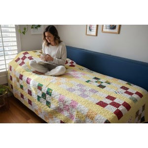 May include: A handmade quilt with a yellow background and a patchwork design featuring squares of red, green, and pink. The quilt is draped over a blue sofa, creating a cozy and inviting scene.