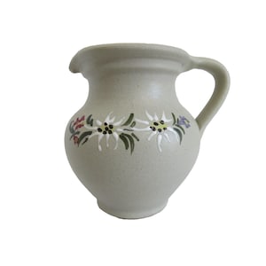 May include: A small, beige ceramic pitcher with a handle and spout. The pitcher is decorated with a floral design featuring white, green, purple, and red flowers. The pitcher has a rounded body and a slightly textured surface.