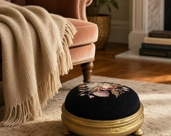 May include: A round footstool with a black fabric top featuring a floral needlepoint design in shades of pink, white, and green. The base is constructed from gold-painted wood with a smooth, curved edge and small, rounded feet for support.