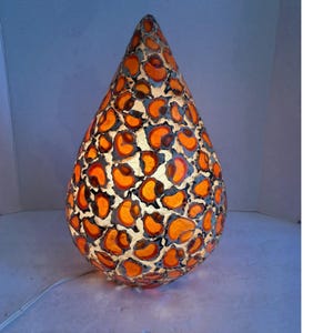 May include: A teardrop-shaped table lamp featuring a translucent glass surface with a repeating orange and black leopard print pattern. The lamp emits a warm glow from within, highlighting the irregular shapes of the spots and the light-colored base.