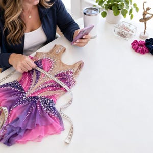 May include: A sleeveless leotard featuring a gradient of pink and purple fabric with a nude-colored mesh bodice. The garment is heavily embellished with clear crystals in a sunburst pattern and includes a ruffled skirt, measured with a tape.