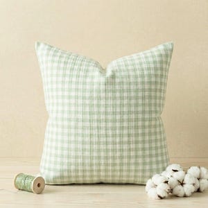 May include: A square throw pillow featuring a light sage green and white gingham check pattern. The fabric has a subtle, crinkled texture. Beside the pillow sits a small wooden spool of green thread and a cluster of white cotton flower bolls.