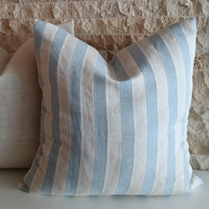 May include: A square pillow with a light blue and white striped pattern. The pillow is made of a soft fabric and has a textured appearance. Another pillow with a neutral color is partially visible in the background. The pillows are likely for home decor.