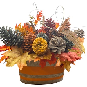 May include: A fall-themed arrangement in a galvanized metal container with an orange band. The arrangement features colorful leaves, pinecones in various colors, and sprigs of wheat. The leaves are in shades of red, orange, and yellow.