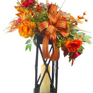 May include: A black metal lantern with a cream-colored candle inside, adorned with a vibrant autumn floral arrangement. The arrangement features orange, red, and green faux flowers, leaves, and berries, accented by a plaid ribbon.