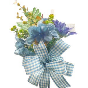 May include: A floral arrangement featuring blue and purple flowers, accented with green foliage and a light blue and white gingham bow. The arrangement is set against a white background.