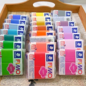 May include: A collection of Fimo Soft modelling clay blocks in various colours, including green, blue, yellow, orange, red, pink, purple, and grey. Each rectangular block is individually wrapped in clear plastic with a white label featuring the Fimo logo.