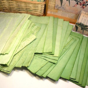 May include: A collection of handmade, green-toned paper sheets in varying shades, arranged in stacks. The paper has a textured appearance, with some sheets showing a lighter, almost yellow-green hue. The papers are likely for crafts or stationery.