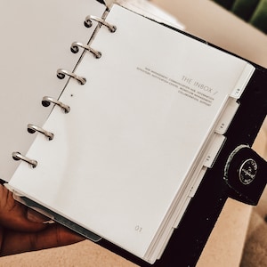 Könnte beinhalten: Ein weißer Planer mit schwarzem Einband und silbernen Ringen. Der Planer hat Registerkarten mit den Beschriftungen "01", "02" und "03". Der Einband des Planers trägt den Text "THE INBOX / TASE MANAGEMENT, COMMUNICATION HUB, INFORMATION REPOSITORY, NOTIFICATION CENTRE, WORKFLOW MANAGEMENT, COLLABORATION, SUPPORT".