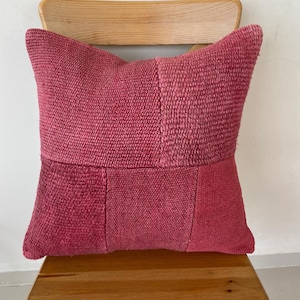 May include: A square pink cushion with a patchwork design. The cushion is made from a textured fabric and has a slightly faded appearance.