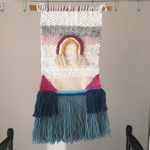 May include: A white woven wall hanging with a rainbow design in pink, orange, yellow, and brown. The hanging has a fringe border in blue and teal.