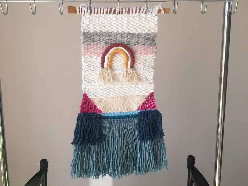 May include: A white woven wall hanging with a rainbow design in pink, orange, yellow, and brown. The hanging has a fringe border in blue and teal.