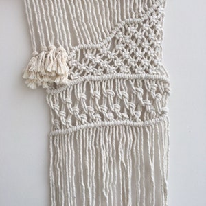 May include: A white macrame wall hanging with a geometric pattern and fringe. The hanging is attached to a wooden dowel.