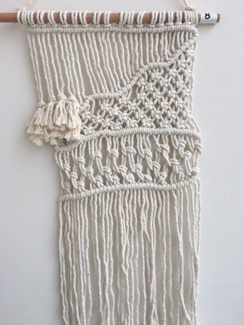 May include: A white macrame wall hanging with a geometric pattern and fringe. The hanging is attached to a wooden dowel.