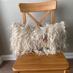 May include: A white fluffy pillow sits on a wooden chair. The pillow is made of a soft, shaggy material and has a slightly irregular shape.