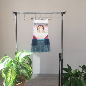 May include: A woven wall hanging with a rainbow design in pink, orange, yellow, and blue. The hanging is made with a variety of textures and colors, including white, gray, and pink. It is hanging on a metal rack with hooks.