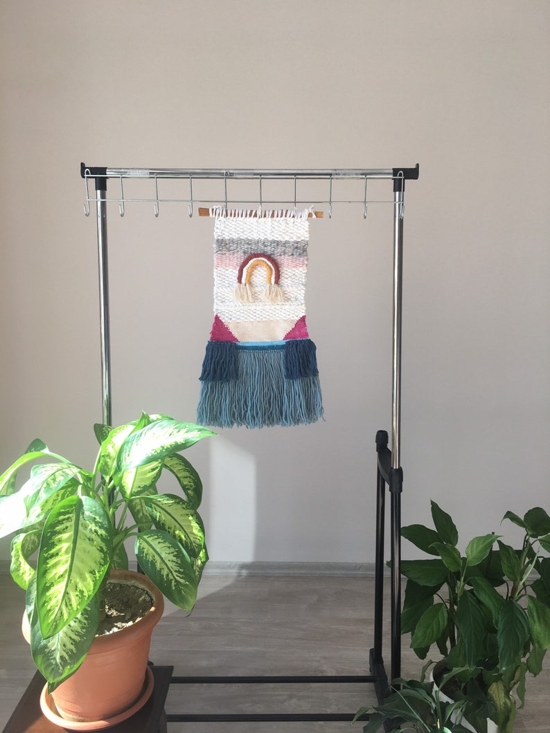 May include: A woven wall hanging with a rainbow design in pink, orange, yellow, and blue. The hanging is made with a variety of textures and colors, including white, gray, and pink. It is hanging on a metal rack with hooks.