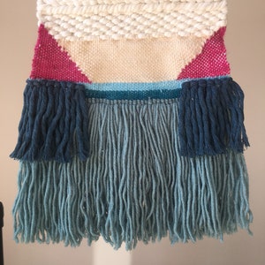 May include: A woven wall hanging with a geometric design in shades of white, pink, blue, and teal. The hanging has a fringe border with long strands of blue yarn.