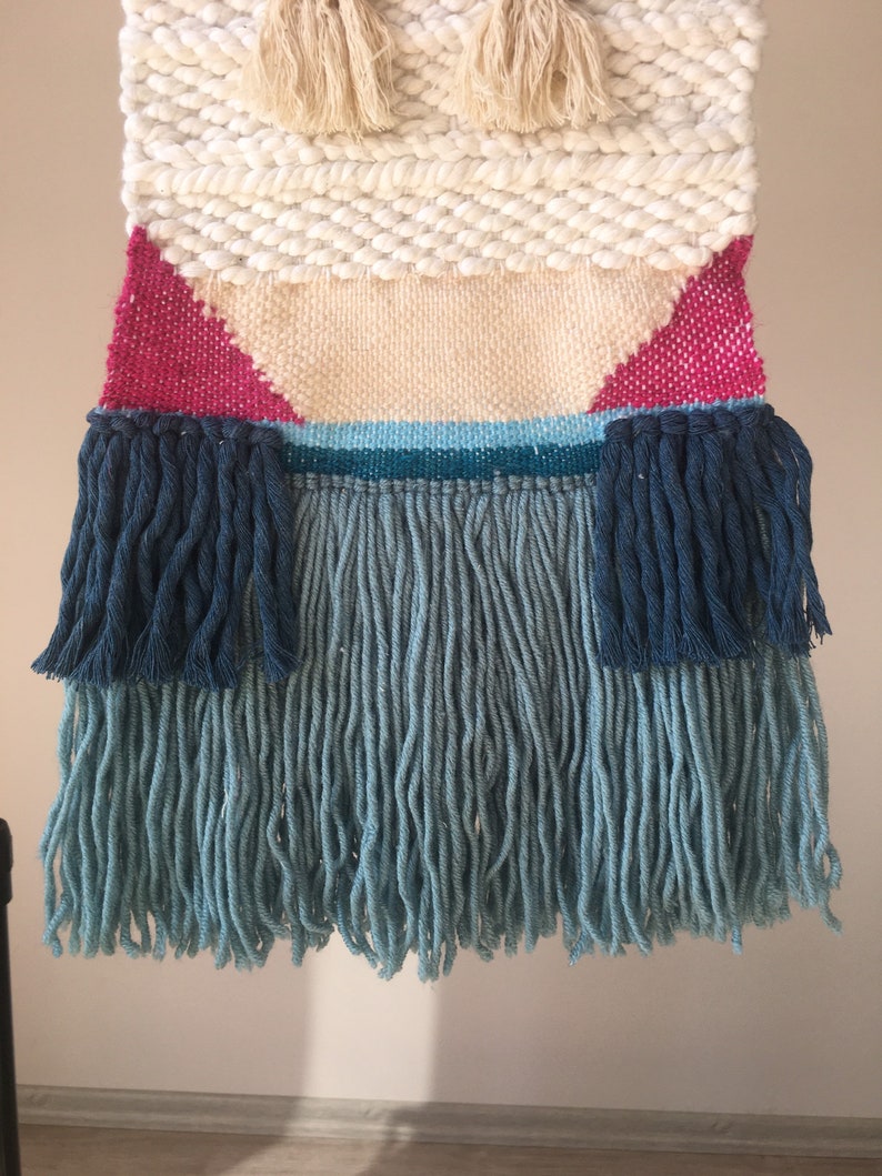 May include: A woven wall hanging with a geometric design in shades of white, pink, blue, and teal. The hanging has a fringe border with long strands of blue yarn.