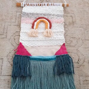 May include: A woven wall hanging with a rainbow design in shades of pink, orange, yellow, and brown. The hanging has a white background and a fringe border in a light blue color.