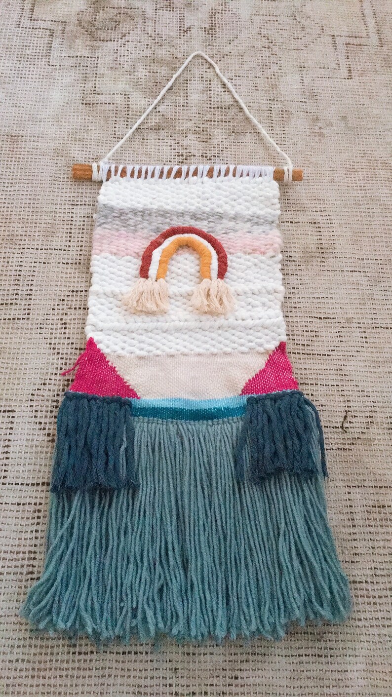 May include: A woven wall hanging with a rainbow design in shades of pink, orange, yellow, and brown. The hanging has a white background and a fringe border in a light blue color.