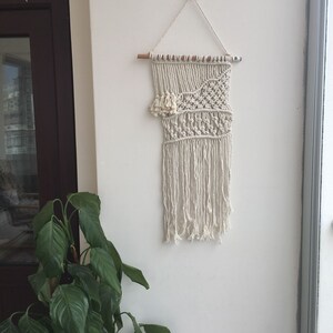 May include: A white macrame wall hanging with a geometric pattern and fringe. The hanging is attached to a wooden dowel rod.