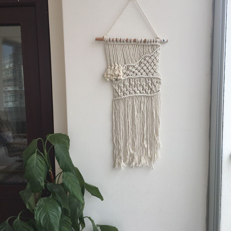 May include: A white macrame wall hanging with a geometric pattern and fringe. The hanging is attached to a wooden dowel rod.