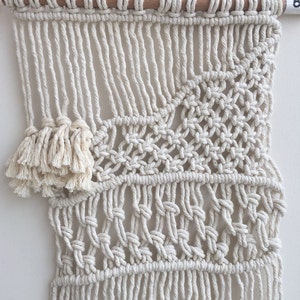 May include: A white macrame wall hanging with a geometric pattern and a fringe border. The hanging is made with natural cotton cord and is attached to a wooden dowel.