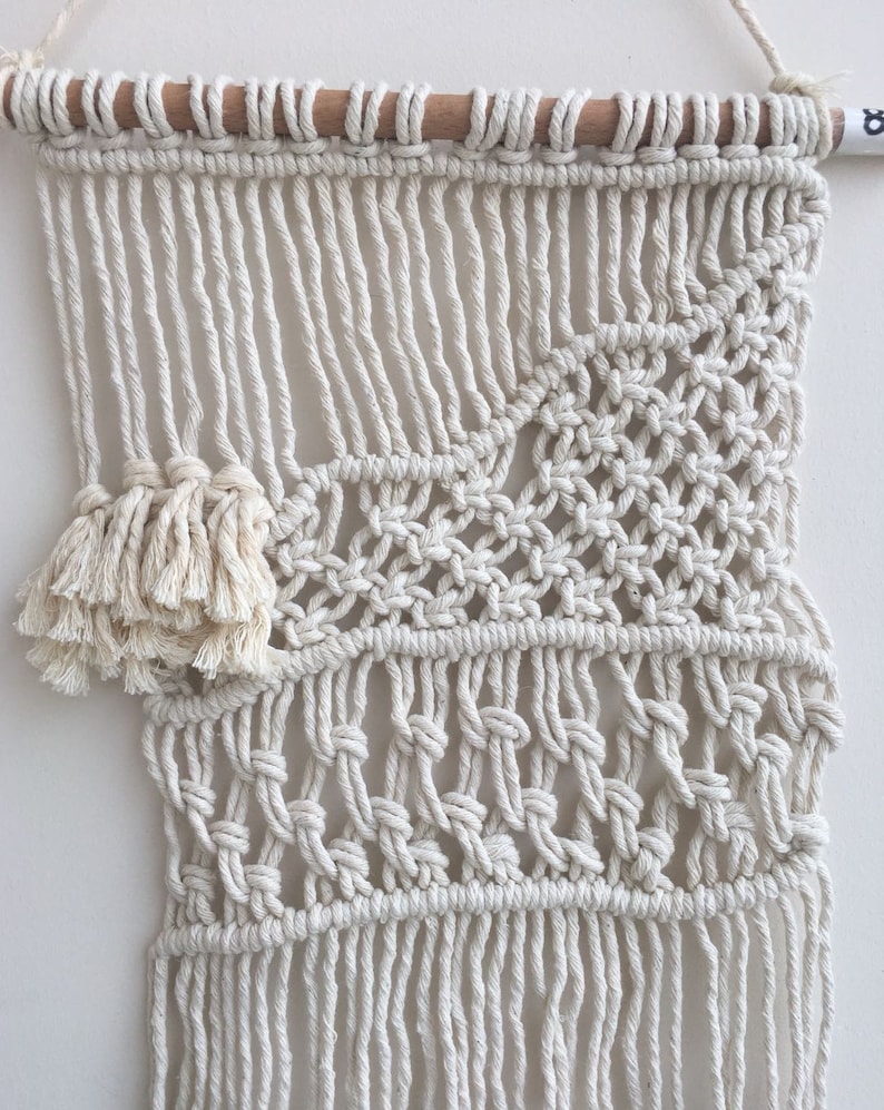 May include: A white macrame wall hanging with a geometric pattern and a fringe border. The hanging is made with natural cotton cord and is attached to a wooden dowel.