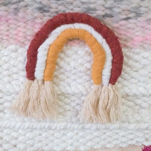 May include: A woven wall hanging with a rainbow design. The rainbow is made of yarn in shades of red, orange, yellow, and white. The background is a white woven fabric with a pink and gray stripe.