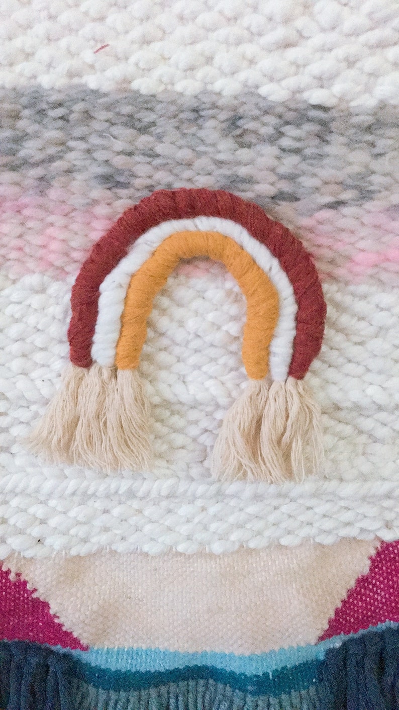 May include: A woven wall hanging with a rainbow design. The rainbow is made of yarn in shades of red, orange, yellow, and white. The background is a white woven fabric with a pink and gray stripe.
