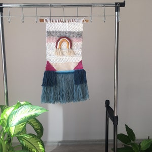 May include: A woven wall hanging with a rainbow design in shades of pink, yellow, orange, and brown. The hanging has a white background with gray and pink stripes. The bottom of the hanging has blue and teal fringe.
