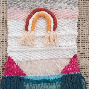 May include: A woven wall hanging with a rainbow design in shades of red, orange, yellow, and white. The hanging has a white background with pink, gray, and blue stripes. The bottom of the hanging has a fringe of blue yarn.