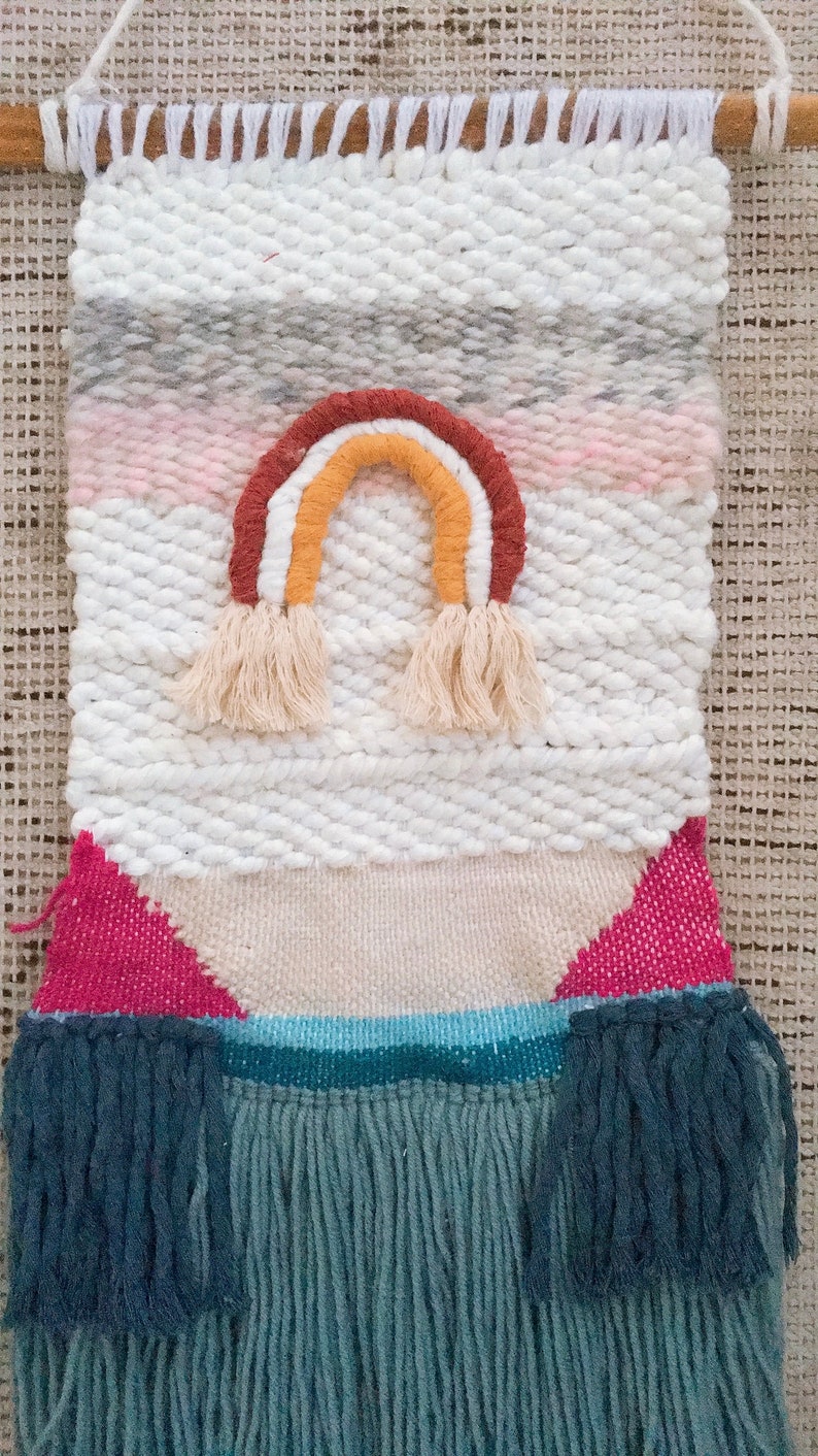 May include: A woven wall hanging with a rainbow design in shades of red, orange, yellow, and white. The hanging has a white background with pink, gray, and blue stripes. The bottom of the hanging has a fringe of blue yarn.