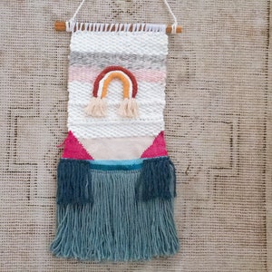 May include: A woven wall hanging with a rainbow design in shades of pink, orange, yellow, and brown. The hanging has a fringe border in a teal color.