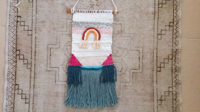 May include: A woven wall hanging with a rainbow design in shades of pink, orange, yellow, and brown. The hanging has a fringe border in a teal color.
