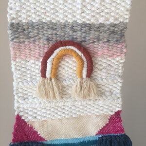 May include: A white woven wall hanging with a rainbow design in shades of red, orange, and yellow. The rainbow has tassels at the bottom. The wall hanging has a pink, gray, and blue striped pattern at the top and a pink, blue, and white geometric pattern at the bottom.