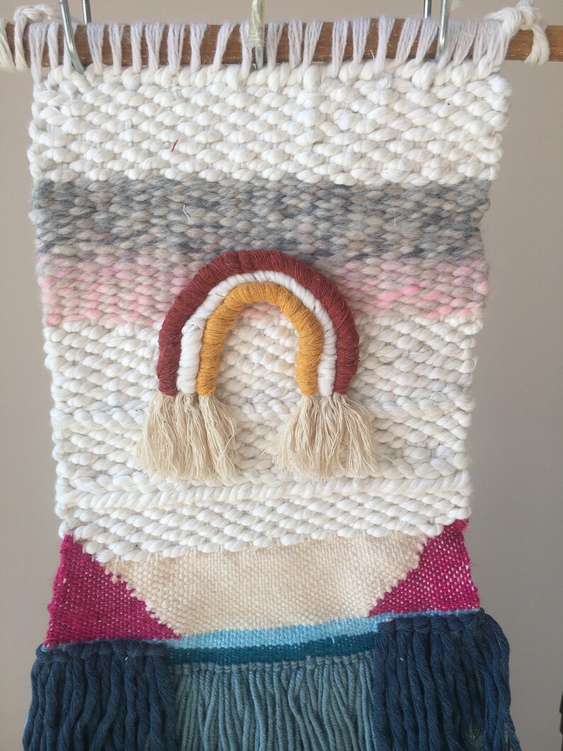 May include: A white woven wall hanging with a rainbow design in shades of red, orange, and yellow. The rainbow has tassels at the bottom. The wall hanging has a pink, gray, and blue striped pattern at the top and a pink, blue, and white geometric pattern at the bottom.