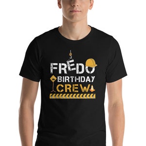 May include: Black t-shirt with the text "FREDO BIRTHDAY CREW" in yellow and black. The text is surrounded by construction-themed graphics, including a hard hat, a traffic cone, and a construction sign.