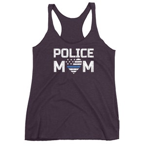 May include: A dark purple racerback tank top featuring the white text POLICE MOM printed on the front. The letter O is replaced by a heart shape containing a stylized American flag design with a single horizontal blue stripe across the center.