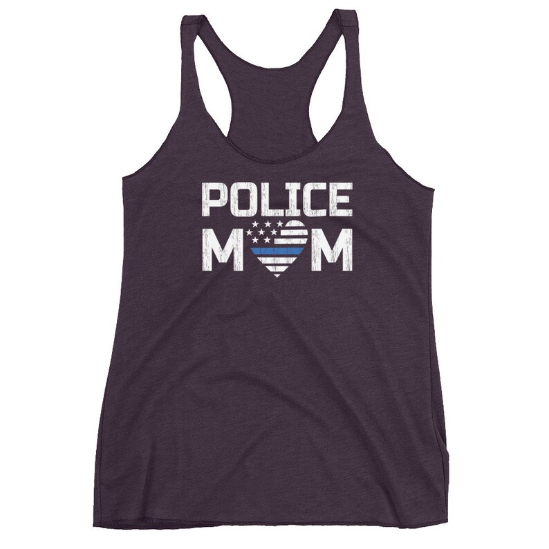 May include: A dark purple racerback tank top featuring the white text POLICE MOM printed on the front. The letter O is replaced by a heart shape containing a stylized American flag design with a single horizontal blue stripe across the center.