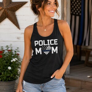 May include: A black sleeveless tank top featuring the white distressed text POLICE MOM with a heart-shaped American flag graphic in the center, which includes a single horizontal blue stripe across the middle of the heart design.